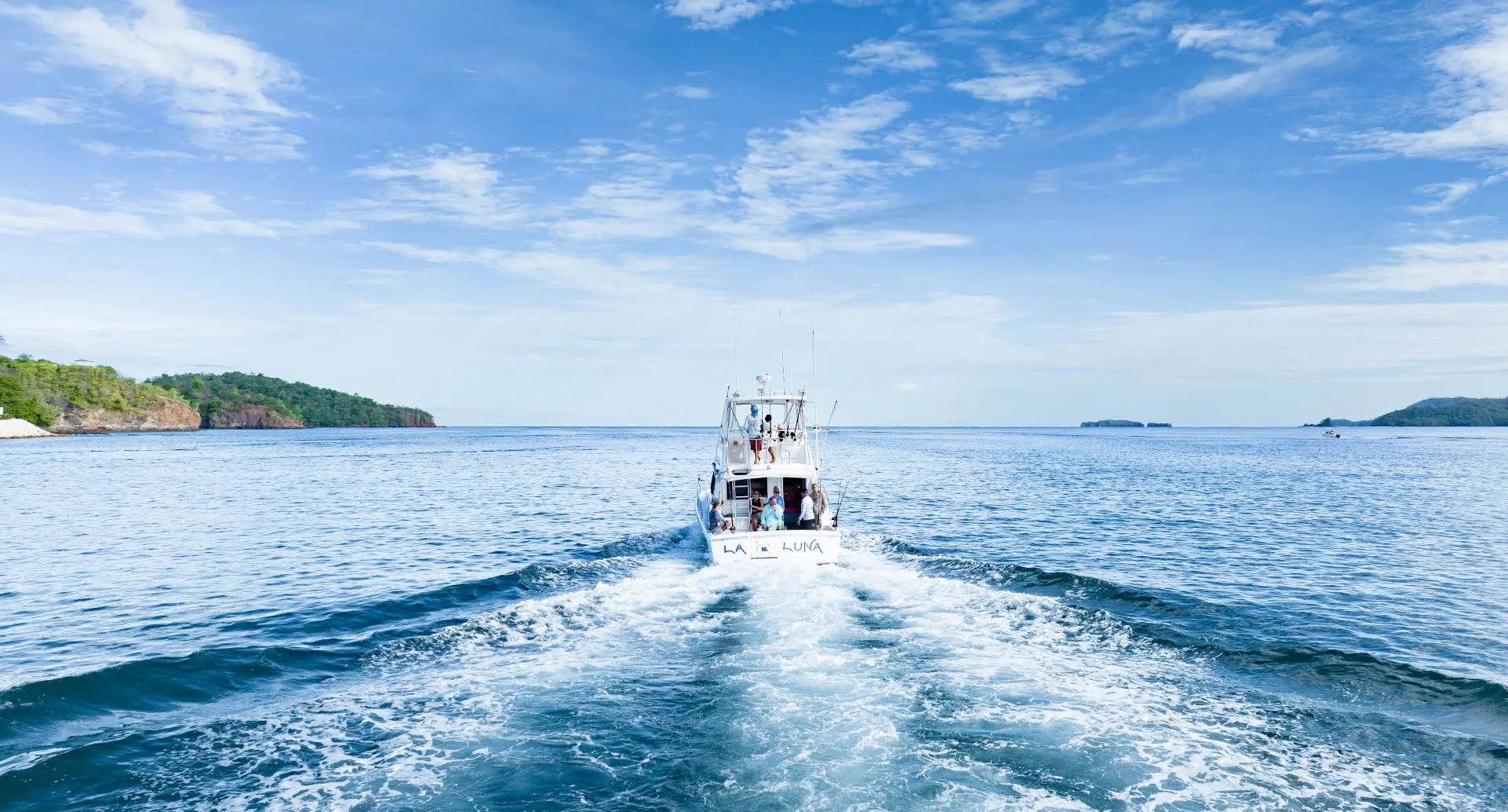 A white motorboat cruises on clear blue water, leaving a trail of foam behind under a clear sky, with distant green islands on the horizon.